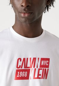 Bela bomba majica iz bombaža z drznim rdečim grafičnim besedilom "CALVIN KLEIN NYC 1968." Klasična zasnova s krožnim izrezom in gladko teksturo.