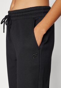 Svarte joggebukser med elastisk midje, snøring og sidelommer. Har en subtil brodert logo på nederste del av benet.