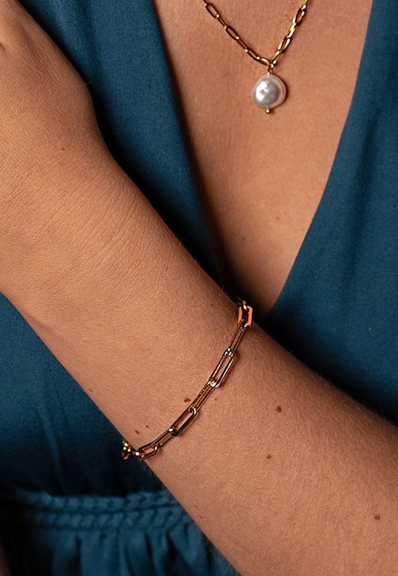 Narukvica u stilu papirnog spajalice zlatne boje, napravljena od metala, s izdužеним karikama, nošena na golom zapešću protiv meke plave tkanine u pozadini.