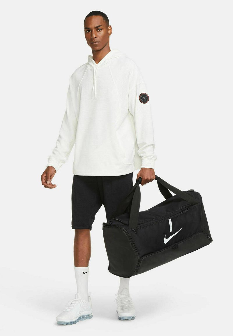 Zwarte Nike sporttas met een groot wit logo, stevige handvatten en een ritsluiting. Draag met een witte hoodie en zwarte shorts.