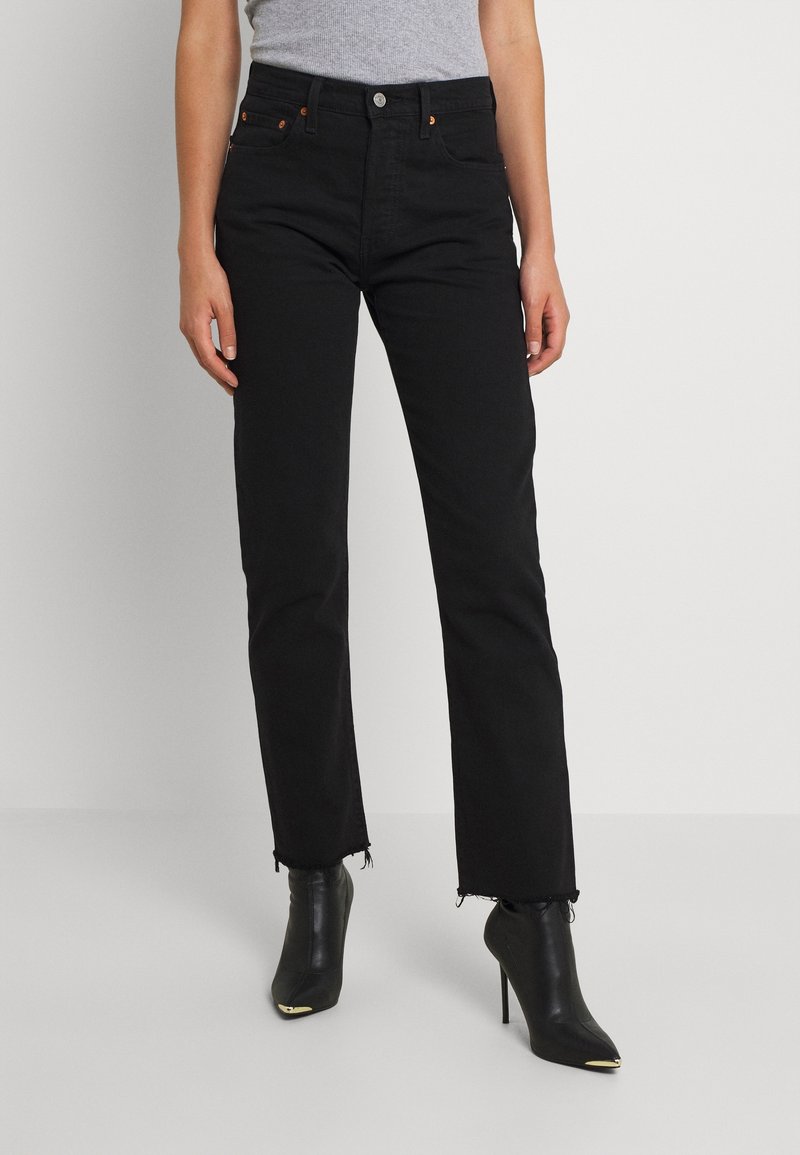 Femme portant un jean droit noir taille haute avec des ourlets effilochés et des bottines noires à talons hauts pointues avec des bouts dorés.