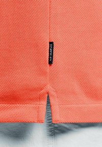 Close-up van koraalstructuurstof met zijsplitdetail en een klein zwart label genaaid in de naad.