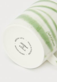 Λευκή κεραμική κούπα με πράσινες λωρίδες και λεία επιφάνεια. Στον πάτο είναι σημειωμένα τα "Matcha Latte", "Iittala" και στοιχεία παραγωγής.