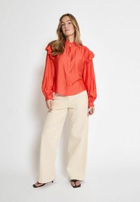 Blusa rossa a maniche lunghe con accenti a volant sulle spalle e sui polsini; abbinata a pantaloni larghi color crema e sandali con tacco a blocco marroni.