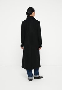 Manteau en laine noir à la coupe longue, col à revers et poignets boutonnés. Il comporte une fente au dos et est associé à un denim retroussé et des chaussures.