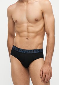 Torse masculin torse nu portant un slip Diesel noir avec une ceinture bleu marine, debout devant un fond clair uni.