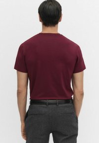 Mand står med ryggen til kameraet, iført en ensfarvet burgunder t-shirt med korteærmer, grå bukser og et sort bælte foran en ensfarvet baggrund.