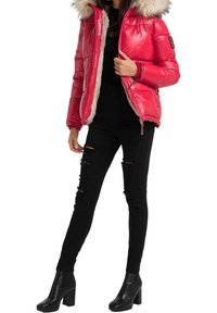 Vrouw in een glanzende rode pufferjack met een bontkap, zwarte gescheurde skinny jeans, een zwarte geribde trui en zwarte enkellaarsjes met hak.