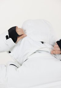 Hvid jakke med sorte detaljer, justerbar hætte, lynlås lukning og elastiske manchetter. Stoffet fremstår glat med isolerende egenskaber.