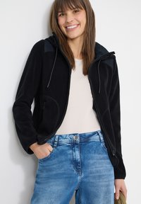 Femme souriante portant un sweat à capuche noir zippé sur un t-shirt blanc et un jean bleu, posant avec une main dans la poche sur un fond blanc.