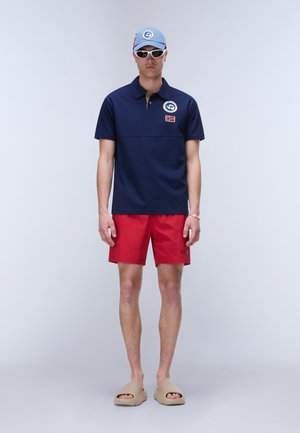 Man draagt een blauw poloshirt met patches, rode short, beige slides, witte zonnebril en blauwe pet, staand tegen een effen achtergrond.