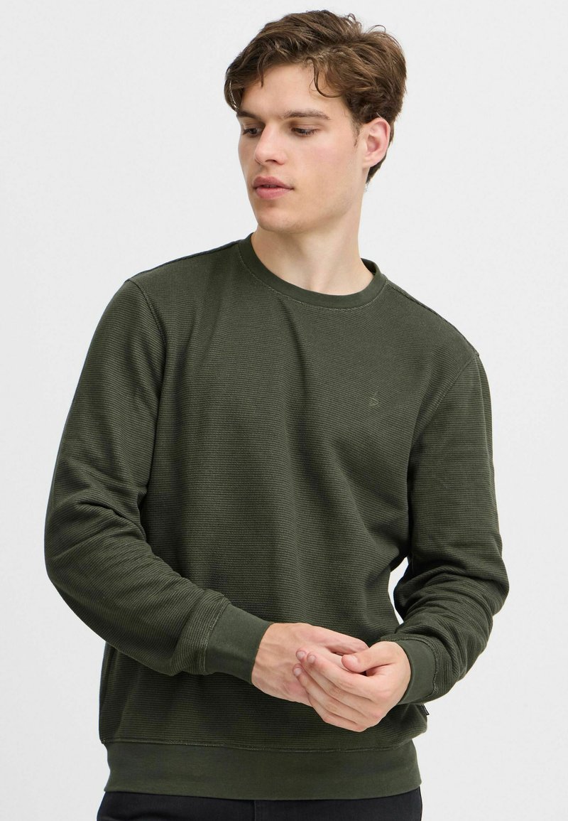 Oliven grøn sweatshirt med ribbet tekstur, rund hals og lange ærmer. Har et diskret logo på brystet samt tætsiddende manchetter og kant.