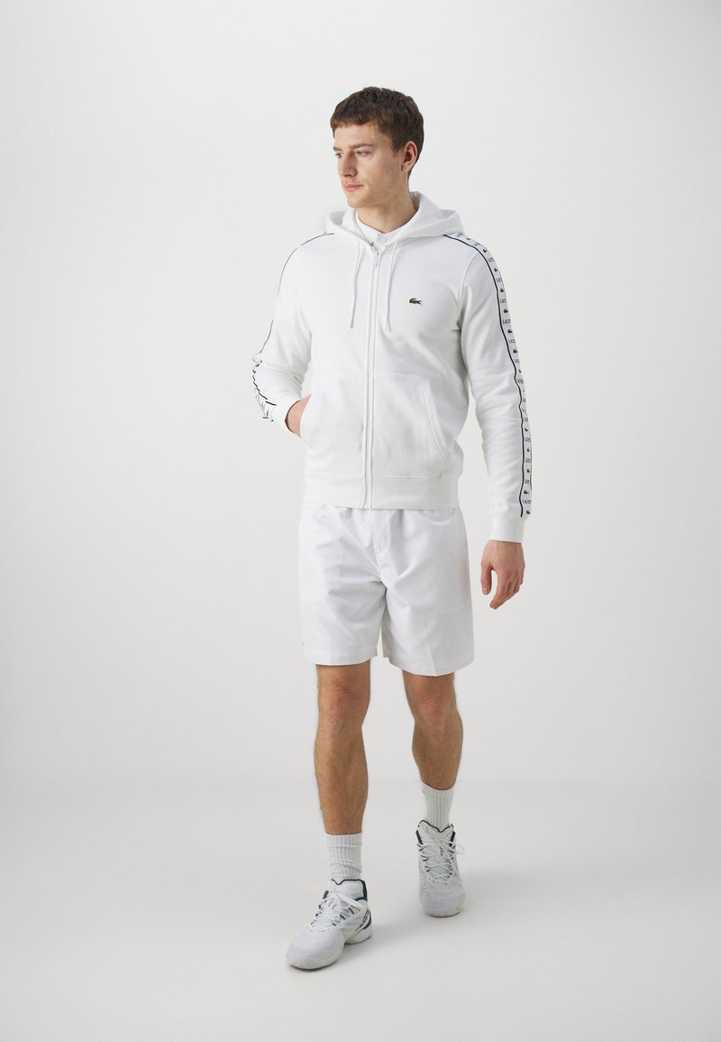 Felpa bianca con zip, decorata con strisce a contrasto nere lungo le maniche, abbinata a pantaloni corti bianchi e sneaker bianche. Tessuto di un morbido effetto.