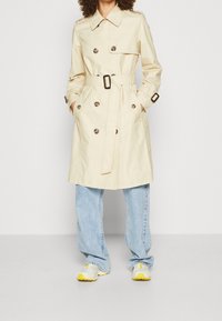 Beige trenssitakki, jossa on kaksiriviset napit, vyötäröllä vyö, sivutaskut ja olkatapit, yhdistettynä löysiin sinisiin farkkuihin ja valkoisiin lenkkareihin.