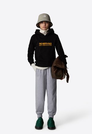 Svart huvtröja med "THE NORTH FACE" logotyp, grå sweatpants, gröna skor och en grå bucket hat. Håller en brun väska. Enkel design.