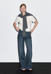 T-shirt bianca, jeans oversized in denim blu scuro e un maglione grigio testurizzato appoggiato sulle spalle. Design semplice con accenti minimi.