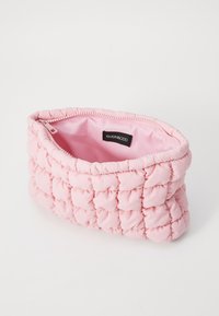 Puffig rosa kosmetikväska med ett kviltat mönster. Har en dragkedjeslutning och en mjuk, slät inredning. Etikett synlig inuti.