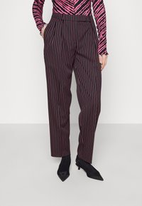 Ušité kalhoty v černé barvě s vertikálními červenými pruhy, s vysokým pasem a bočními kapsami, zkombinované s špičatými černými podpatky.