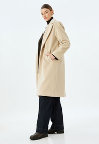 Beige uldblandingsfrakke med reverskrave, sidelommer og lange ærmer. Båret over en sort turtleneck og paired med mørke denim og støvler.