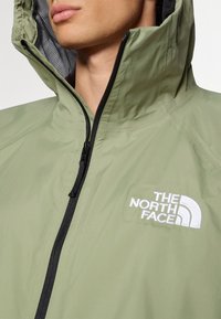 Gros plan d'une personne portant une veste à capuche vert clair avec une fermeture éclair noire et le logo blanc The North Face sur la poitrine.
