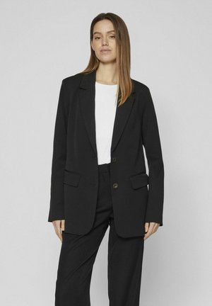 Blazer nero con una vestibilità sartoriale, due tasche frontali e chiusura a un bottone. Indossato sopra una maglietta bianca, con tessuto testurizzato.