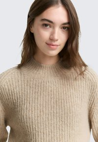 Beige gestrickter Pullover mit hohem Kragen, strukturiertem Muster und gerippten Bündchen. Weiches Material. Enganliegendes Design mit entspannter Silhouette.
