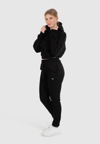 Sudadera negra recortada con mangas largas y cremallera, combinada con joggers negros con bolsillos laterales. Zapatillas blancas completan el look.