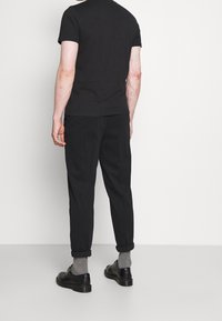 Camiseta negra de manga corta, pantalones negros con puños doblados, calcetines grises y zapatos negros; todo presentado desde una vista trasera sobre un fondo blanco.
