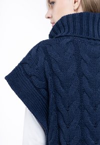 Marineblå strikket poncho med høj krave, der har et struktureret kabelstrik mønster og korte ærmer, lavet af tykt garn.
