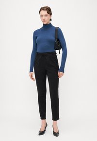 Jersey de cuello alto azul, pantalones negros ajustados y tacones altos negros. Diseño estructurado, textura suave y detalle de cordón en los pantalones.