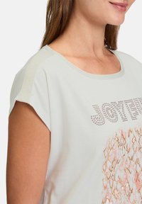 Jasnoszara bluzka z krótkim rękawem z okrągłym dekoltem, błyszczącym napisem oraz teksturowanym wzorem z metalicznymi akcentami w kolorze różowym i złotym.