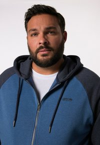 Blauwe zip-up hoodie met donkerblauwe accenten, voorzien van een capuchon met trekkoord en een logo op de borst. Zacht materiaal en ribgebreide manchetten.