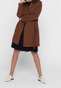 Una mujer se encuentra de pie con las piernas separadas, vistiendo un abrigo marrón sobre un vestido negro y zapatos blancos sin cordones, frente a un fondo claro y sencillo.