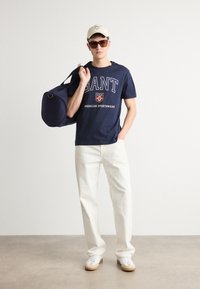 Tricou bleumarin cu logo-ul alb "GANT", pantaloni de culoare deschisă, adidași albi și o șapcă bej. Poartă o geantă bleumarin.