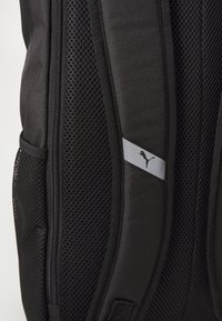 Crna ranac s mrežastim detaljima, s glatkom površinom tkanine i srebrnim Puma logom na sivoj pruzi. Bočna džep vidljiv.