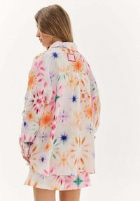 Žena oblečená v pestrofarebnej tričku s dlhým rukávom v štýle tie-dye a zodpovedajúcej sukni, stojaca chrbtom k fotoaparátu a pozerajúca sa na stranu.