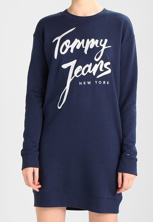 Długa granatowa sukienka dresowa z białym napisem „Tommy Jeans New York” z przodu, noszona przez osobę na jednolitym tle.