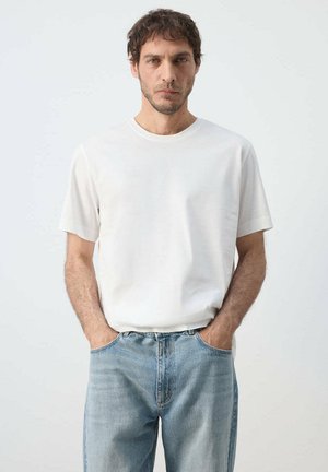 Mand står mod en ensfarvet baggrund iført en hvid T-shirt og blå jeans, med hænderne i lommerne og ansigtet vendt fremad med et neutralt udtryk.