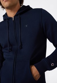 Uomo in felpa blu navy con zip e coulisse, mano nella tasca anteriore, piccolo logo sul petto e sul polsino della manica.