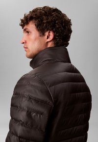 Pruun tikitud puffer-jakk kõrge kraega, nähtavate õmblustega ja siledapinnaline, küljepealt vaadatuna.