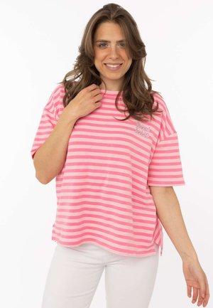Lächelnde Frau mit langen braunen Haaren, die ein lockeres rosa-weiß gestreiftes T-Shirt und weiße Hosen trägt und ihren Hemdkragen berührt.