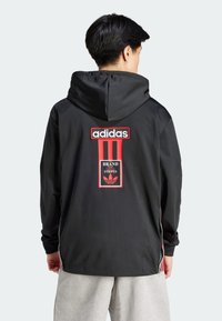 Giacca nera con cappuccio caratterizzata da accenti rossi, tre strisce verticali e un logo Adidas sul retro. Il materiale sembra liscio e leggero.