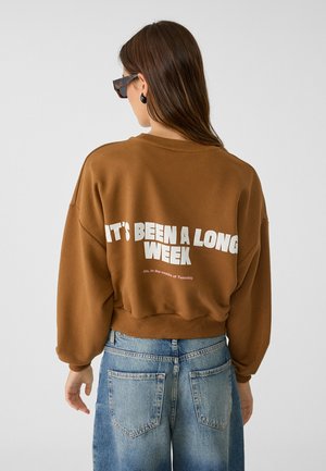 Femme aux cheveux longs portant des lunettes de soleil écaille de tortue, un sweat-shirt marron avec le texte « IT'S BEEN A LONG WEEK » et un jean bleu, vue de dos.