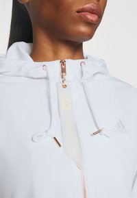 Vit zip-up jacka med huva, försedd med en roséguldig dragkedja, dragsko och en broderad logotyp. Slät tygtextur.