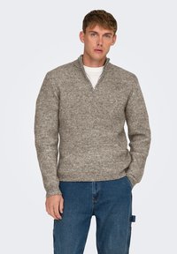 Lichtgrijze geribbelde trui met een halve rits kraag en lange mouwen, gemaakt van gestructureerd breimateriaal. Gecombineerd met blauwe denim jeans.