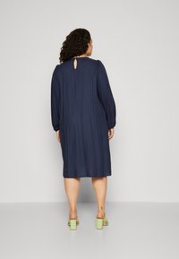 Tamsiai mėlyna suknelė su ilgomis pūstomis rankovėmis, raktine nugaros dalimi ir laisvu siluetu, derinama su šviesiai žaliomis blokiniais kulnais.