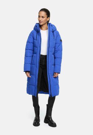 Lange pufferjas in helderblauw, met een capuchon, ritsluiting, ruitpatroon en zwarte accenten. Gepaard met zwarte broeken en chunky laarzen.