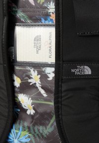 Black fabric pouch with a floral interior pattern featuring white daisies and greenery. Includes a card labeled "Flora Alpina" and brand logos.