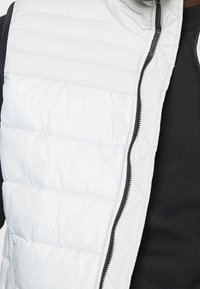 Vit kviltad väst med hög krage, prydd med en svart dragkedja och en slät textur, lagd över en svart tröja.