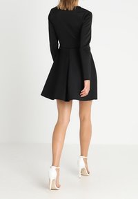 Musta pitkähihaiset puku, jossa on istuva yläosa ja liehuva helma, valmistettu sileästä kankaasta. Päällä valkoiset korkokengät.